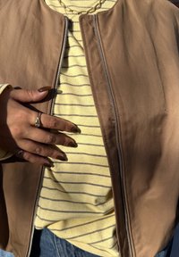 Brun bomberjakke med glidelås, båret over en gul stripete t-skjorte. Har en kjedekjede; negler malt mørke med ringer på fingrene.