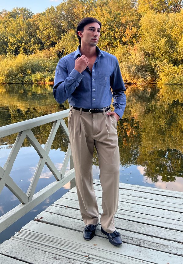Jeune homme portant une chemise bleue et un pantalon beige se tient sur un quai en bois au bord d’un lac calme avec des arbres d’automne en arrière-plan.