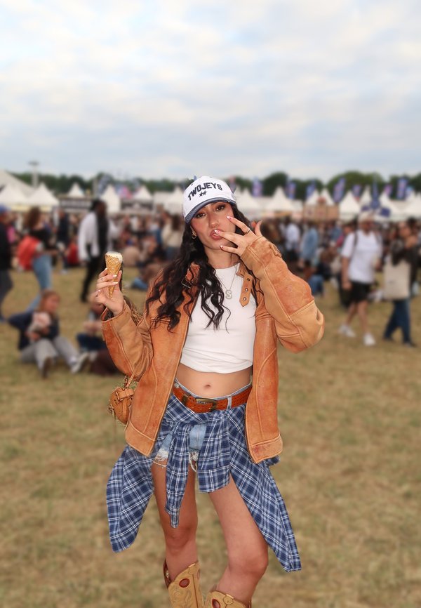 Giovane donna con cappello e giacca marrone che mangia un gelato a un festival all'aperto con persone e tende sullo sfondo.