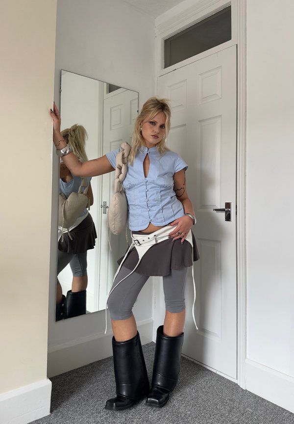 Junge Person mit blonden Haaren steht neben einem Spiegel in einem Raum und trägt ein blaues Oberteil, graue Leggings, schwarze Stiefel und einen grauen Rock mit weißen Trägern.