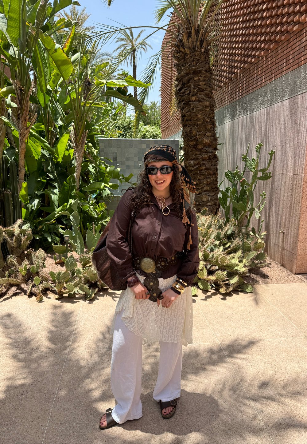 Camisa castanha de mangas longas, saia branca texturada e calças brancas largas. Acessórios: óculos de sol oversized, lenço de cabeça estampado e um cinto grosso.