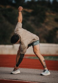Silhouette athlétique s'étirant sur une piste ; portant un t-shirt beige clair à motif, un short vert et des chaussures de course grises avec des accents.