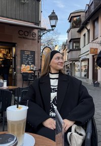 Manteau noir oversize avec un pull blanc orné de motifs de montagnes. Latte avec mousse sur une table en bois, café en milieu urbain.