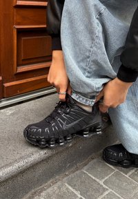 Personne portant un jean bleu clair et des baskets noires accroupie sur une marche en pierre grise près d'une porte en bois.