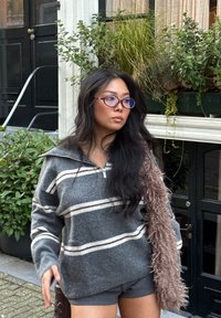 Grijs gestreepte trui met een ritssluiting bij de kraag, gecombineerd met gestructureerde korte mouwen en een zacht roze accessoire. Buitenlocatie met groen op de achtergrond.