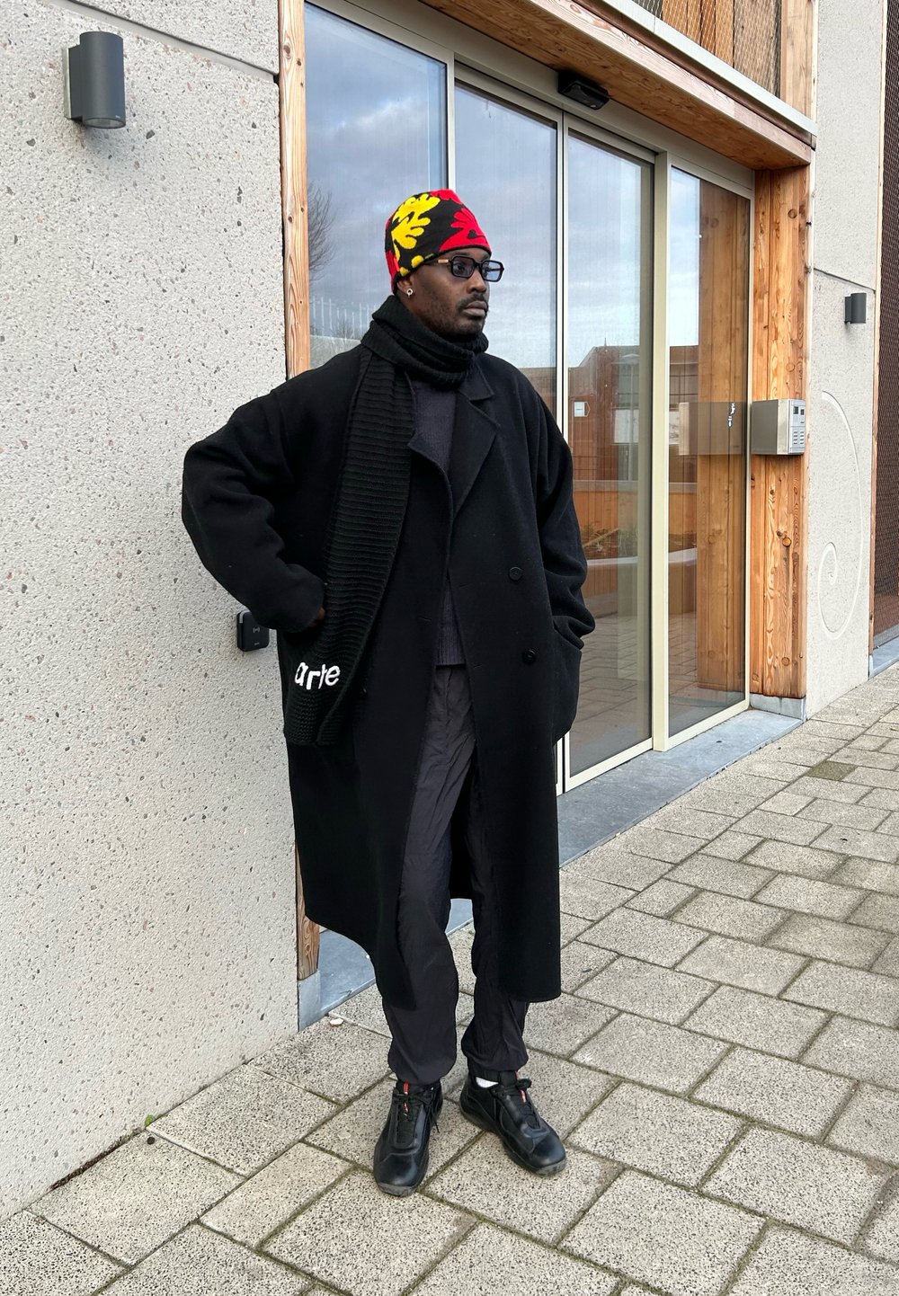 Um longo casaco preto feito de lã, com um cachecol preto texturizado, calças pretas e ténis pretos. A pessoa usa um gorro colorido com um padrão.