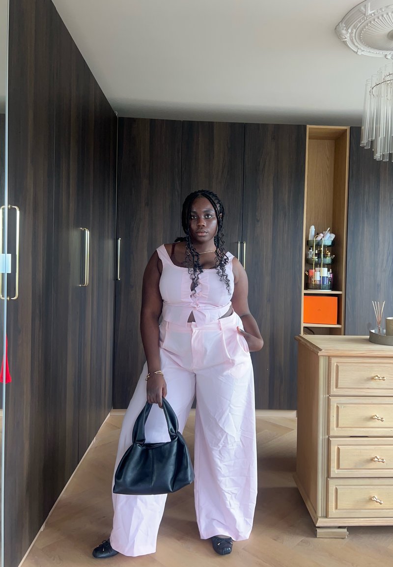 Femme en haut sans manches rose clair et pantalon large tenant un sac à main noir se tient dans une pièce avec des armoires en bois foncé et des tiroirs en bois clair.
