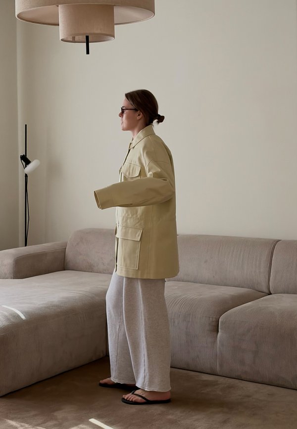 Vrouw met een beige jas en grijze wijde broek staat bij een lichtbruine bank in een minimalistische woonkamer met een vloerlamp en plafondlamp.