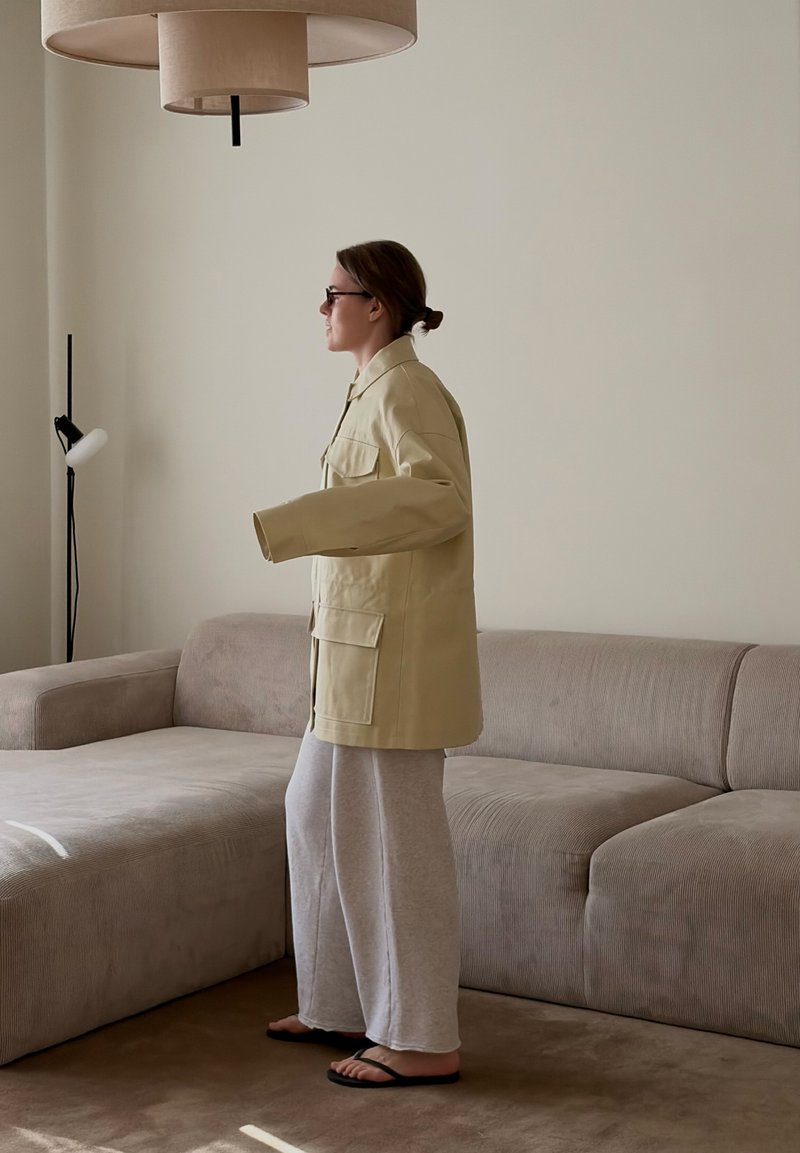 Mujer vestida con chaqueta beige y pantalones anchos grises, de pie junto a un sofá marrón claro en una sala de estar minimalista con lámpara de pie y luz de techo.