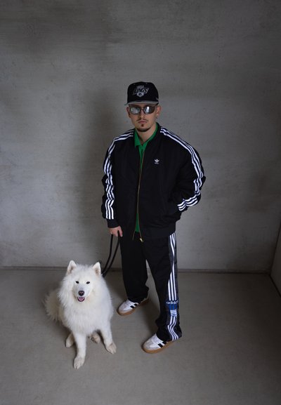 Chaqueta negra con cremallera y rayas blancas, polo verde, pantalones negros y zapatillas blancas. Un perro blanco y esponjoso se sienta al lado de la persona.