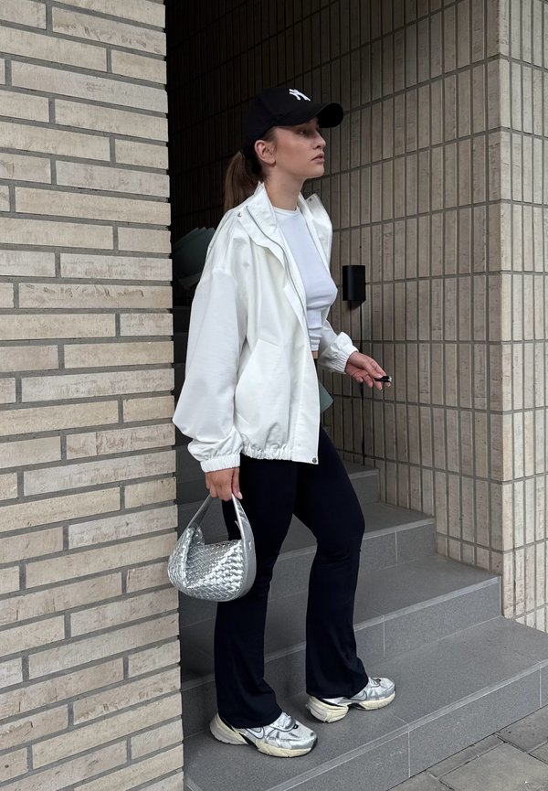 Ung kvinde iført hvid jakke, sort kasket, sorte bukser og sølvfarvede sneakers står på grå trapper og holder en sølvfarvet flettet håndtaske.