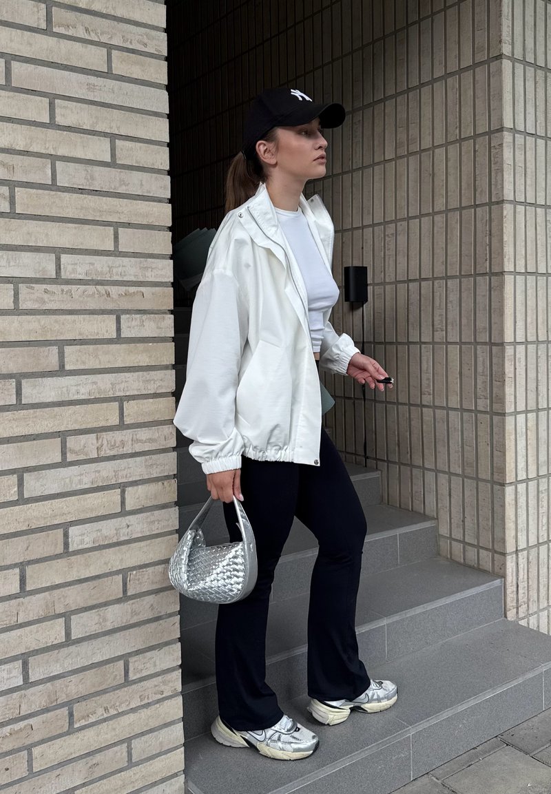 Jeune femme portant une veste blanche, une casquette noire, un pantalon noir et des baskets argentées, debout sur des escaliers gris tenant un sac à main tissé argenté.