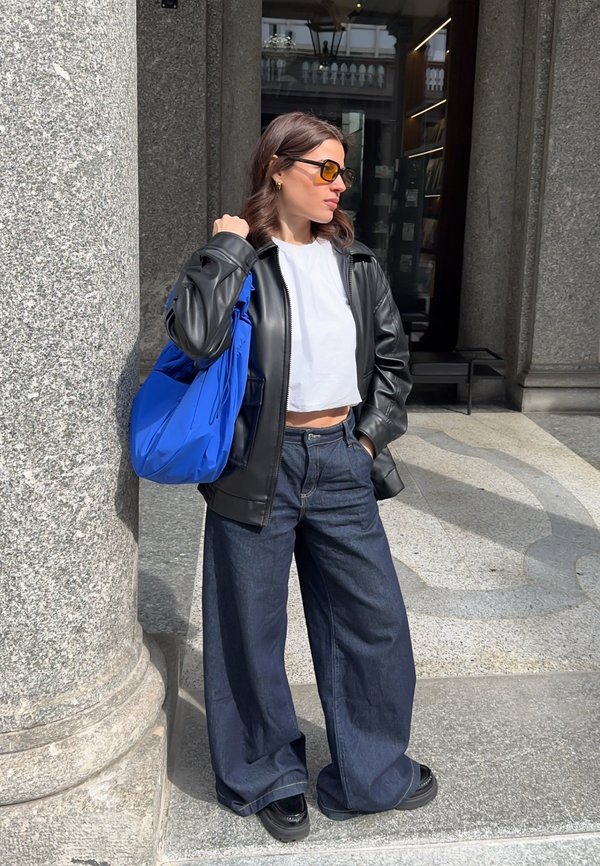 Frau mit Sonnenbrille, schwarze Lederjacke, weißes bauchfreies Top, weite dunkle Jeans, hält eine leuchtend blaue Tasche, steht neben einer Steinsäule im Freien.