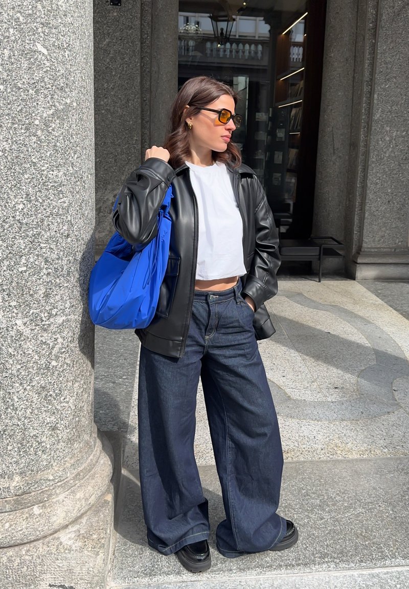 Frau mit Sonnenbrille, schwarze Lederjacke, weißes bauchfreies Top, weite dunkle Jeans, hält eine leuchtend blaue Tasche, steht neben einer Steinsäule im Freien.