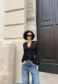 Kvinne med krøllet hår iført svart knappet topp, store solbriller og blå jeans står foran en beige steinvegg nær en stor mørk tredør.