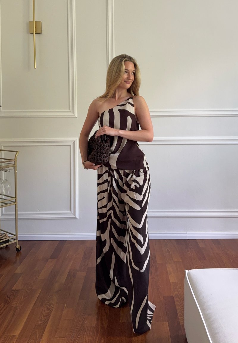 Femme portant un top asymétrique à motif abstrait noir et blanc et un pantalon large, tenant une pochette noire texturée dans une pièce lumineuse et minimaliste.