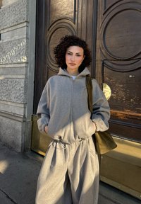 Kvinde med krøllet hår iført lysegrå hættetrøje med lynlås og matchende brede bukser, stående med hænderne i lommerne ved en trædør med messingdetaljer.