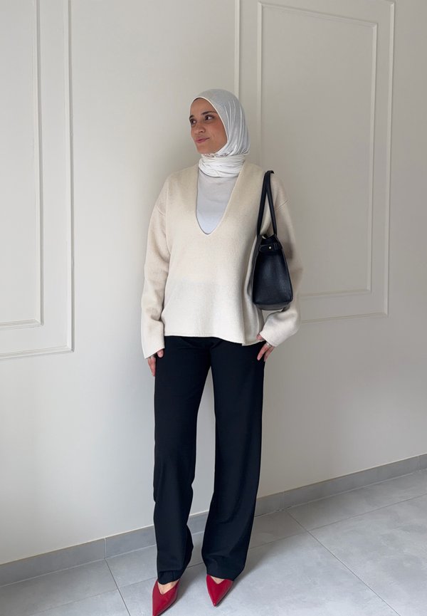 Femme en hijab blanc et pull, pantalon noir, chaussures rouges et sac à épaule noir, debout devant un mur blanc uni à l'intérieur.