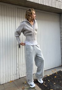 Lys grå kort strikkegenser med ribbestruktur, kombinert med grå joggebukse. Modellen står foran en teksturert vegg med et subtilt mursteinsmønster.