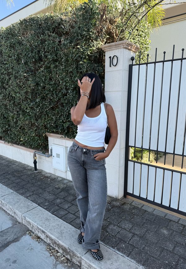 Top branco sem mangas, jeans de ganga soltos cinzentos, sapatos baixos pretos com bolinhas. De pé ao lado de uma coluna branca marcada com '10' e folhagem verde ao fundo.