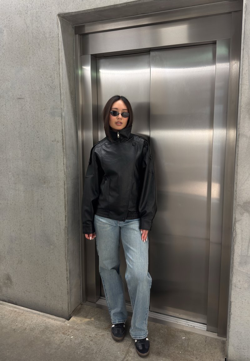 Woman wearing black leather jacket, sunglasses, light blue jeans, and sneakers leaning against closed stainless steel elevator doors.