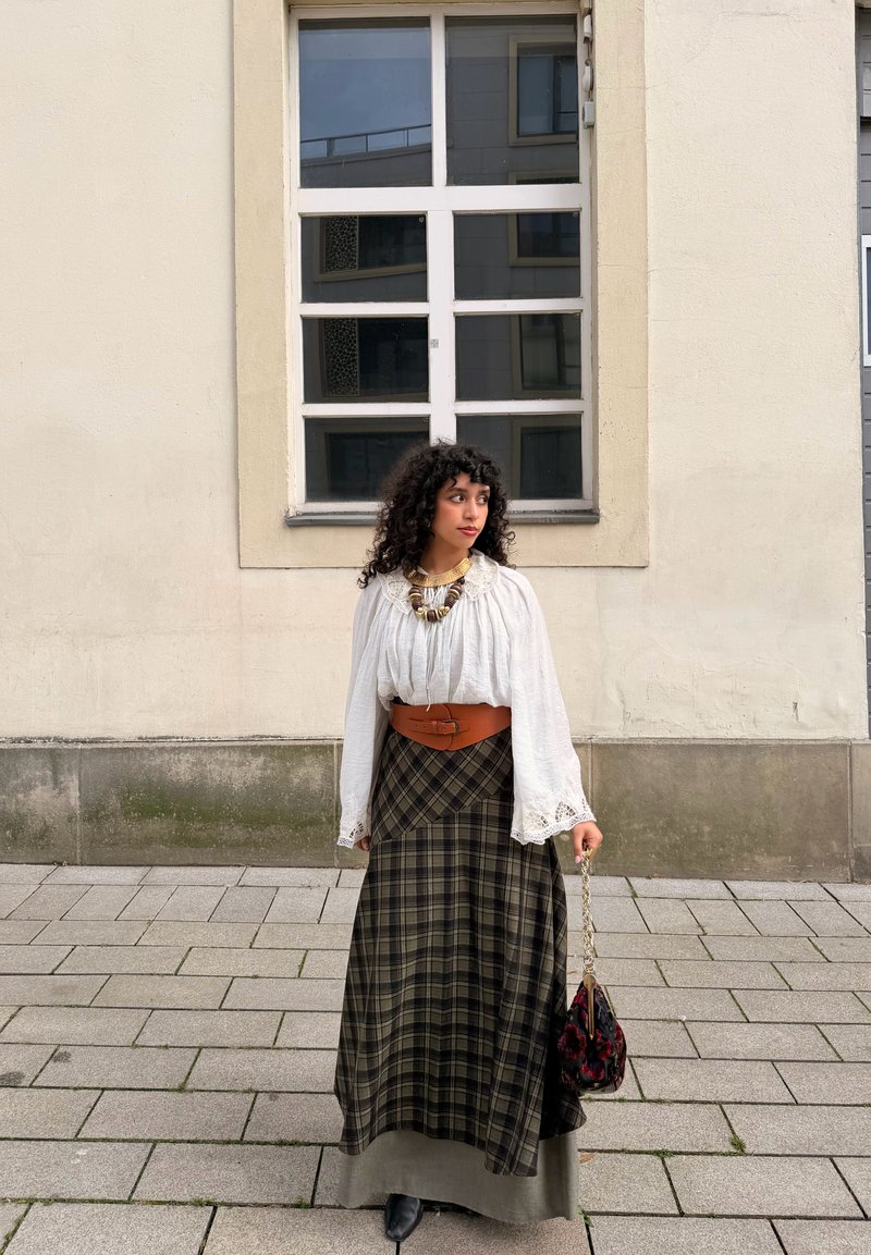 Kvinna med lockigt hår, klädd i vit blus, stort halsband, brett brunt bälte, rutig kjol och håller en liten handväska, stående på en kaklad trottoar.