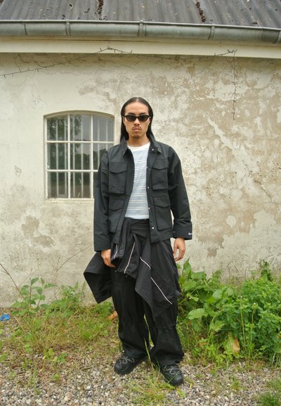 Chaqueta negra con múltiples bolsillos, camisa blanca de rayas debajo, pantalones negros holgados y gafas de sol oscuras que lleva puesta una persona de pie al aire libre.