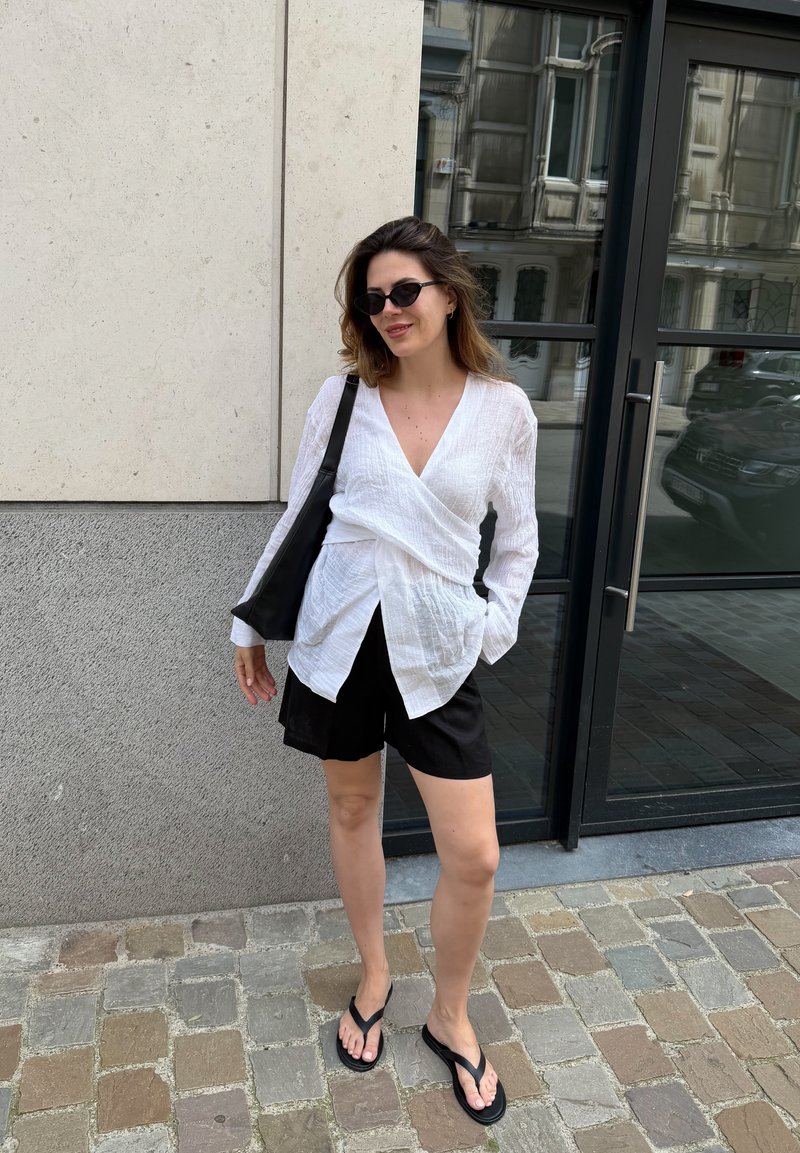 Frau trägt weiße Wickelbluse, schwarze Shorts, Flip-Flops und Sonnenbrille, steht auf Kopfsteinpflaster nahe Glastür und Steinmauer.