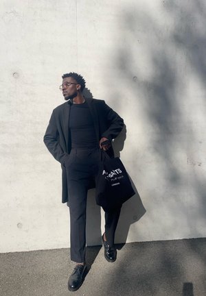 Abrigo negro sobre una blusa negra ajustada, pantalones oscuros y zapatos negros. Sosteniendo una bolsa tote negra con texto blanco. Fondo de concreto.