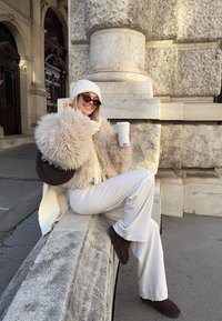 Vaalean värinen turkistakki tummilla yksityiskohdilla, beige leveälahkeiset housut ja ruskeat saappaat. Pitää kädessään ruudullista mukia ja istuu kivisellä reunalla.