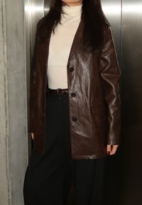 Άτομο που φοράει καφέ δερμάτινο σακάκι πάνω από κρεμ ζιβάγκο και μαύρο παντελόνι, στέκεται μπροστά σε μεταλλική πόρτα ανελκυστήρα και τσιμεντένιο τοίχο.