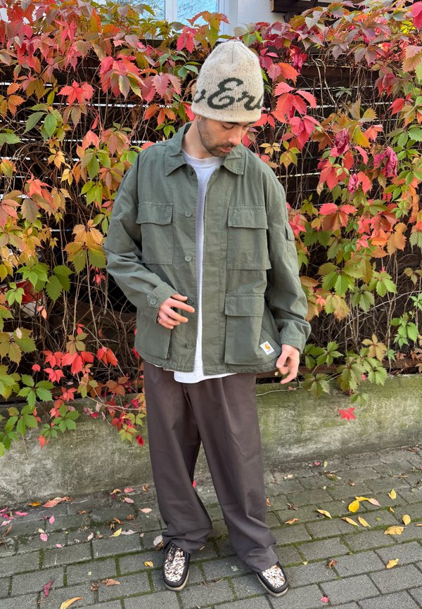 Homme portant une veste verte, un bonnet beige, un pantalon gris et des chaussures à motifs, debout sur un sol pavé avec des feuilles rouges et vertes sur une clôture derrière.