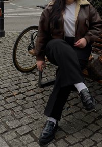 Brun læder bomberjakke med en beige shearlingkrave, hvid t-shirt, sorte vidde bukser og sorte loafers med hvide sokker.