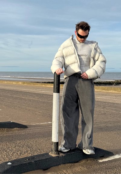 Chaqueta de plumas blanca, camiseta gráfica gris y pantalones grises holgados. De pie junto a un poste en blanco y negro sobre una superficie arenosa cerca de la costa.