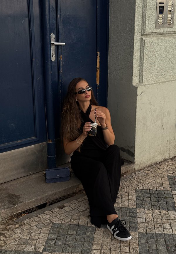Una mujer con gafas de sol negras y ropa negra está sentada junto a una puerta azul, sosteniendo una bebida en una calle empedrada.