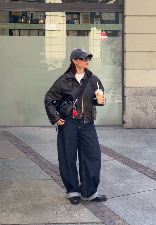 Mujer con chaqueta de cuero oscura y vaqueros anchos sosteniendo un café con hielo, de pie en una acera de la ciudad cerca de una ventana de vidrio.
