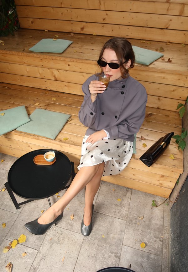 Femme portant des lunettes de soleil et une veste grise, elle boit dans un verre assise sur des marches en bois à côté d'une petite table noire avec une tasse.