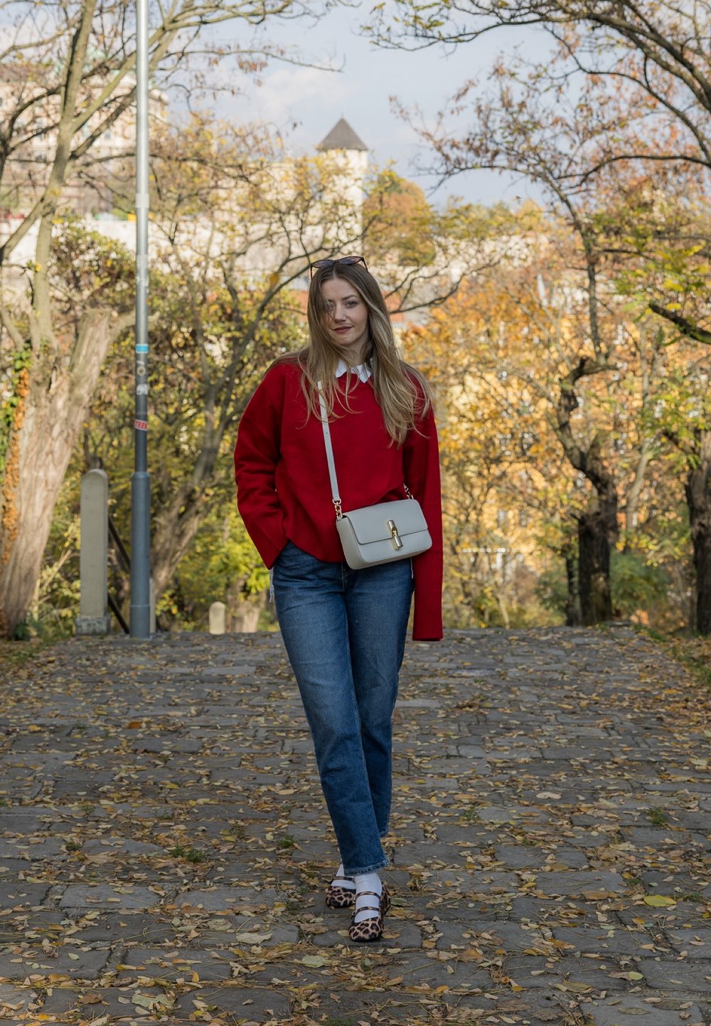 Nő piros pulóverben, kék farmerben, leopárdmintás cipőben sétál falevelekkel borított macskaköves ösvényen, bézs oldaltáska a vállán, mögötte őszi fák.