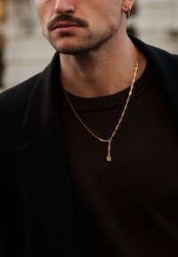 Collier chaîne en or avec maillons et pendentif ; porté sur un pull côtelé brun foncé, accompagné d'un manteau noir.