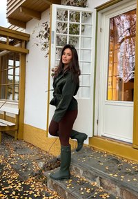 Donna con giacca verde scuro e stivali apre una porta bianca di una casa con rifiniture gialle, con foglie autunnali sui gradini e sul terreno di pietra.