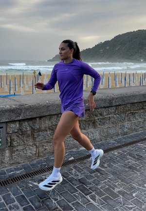 Corridore in una maglietta a maniche lunghe viola e pantaloni corti coordinati, che indossa scarpe da ginnastica bianche con dettagli neri, mentre corre su un sentiero in pietra vicino alla spiaggia.