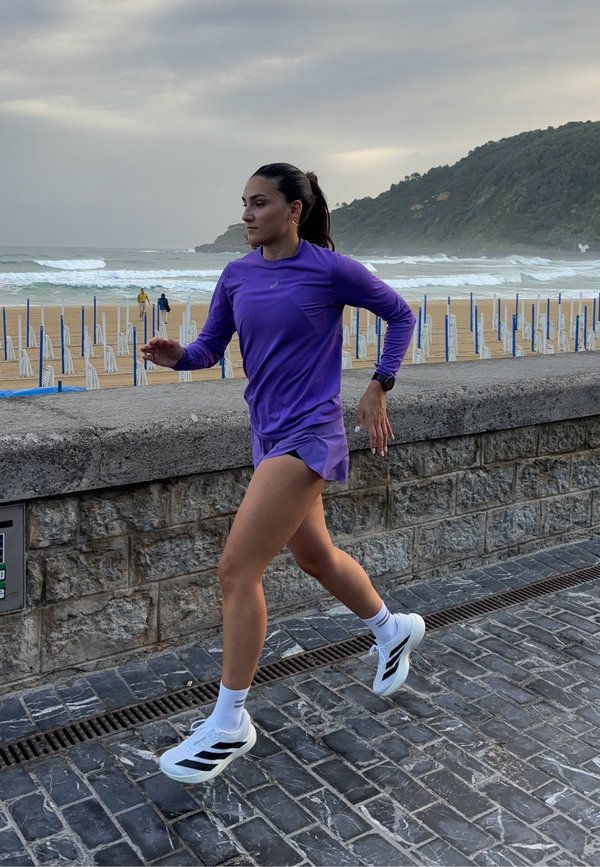 Corredor com uma camisa de manga longa roxa e calções combinando, usando sapatilhas brancas com detalhes pretos, a correr numa passadeira de pedras à beira-mar.