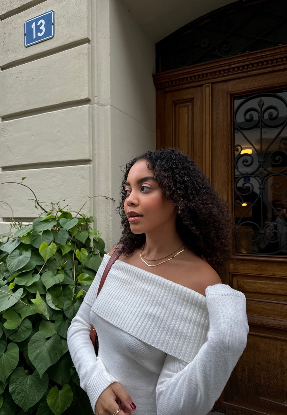 Pull blanc à épaules dénudées avec texture côtelée, manches cintrées, collier doré à plusieurs couches et sac à bandoulière marron, entouré de feuillage vert.