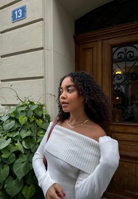 Pull blanc à épaules dénudées avec texture côtelée, manches cintrées, collier doré à plusieurs couches et sac à bandoulière marron, entouré de feuillage vert.