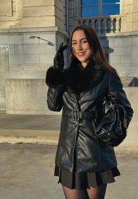 Manteau en cuir noir avec un col et des poignets en fourrure, ceinturé à la taille, porté sur une jupe plissée noire. Sac à main brillant à la main. Lumière chaleureuse du soleil couchant.