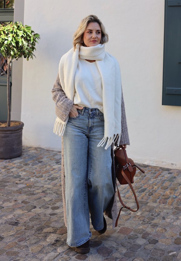 Une femme marche dans une rue pavée, vêtue d’une écharpe blanche, d’un pull et d’un jean à jambes larges, et portant un sac à main en cuir marron.