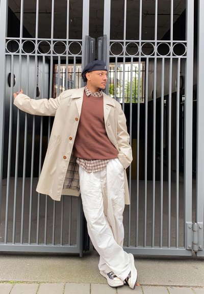 Gabardina beige con grandes botones sobre un suéter marrón y una camisa a cuadros. Pantalones blancos, zapatillas oscuras, de pie contra puertas de metal.