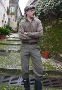 Homme portant une casquette marron, un pull beige, un pantalon gris et des bottes de pluie noires, debout les bras croisés dans une ruelle pavée couverte de mousse.