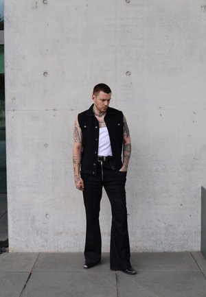 Chaleco de mezclilla negro sobre una camiseta blanca de tirantes, combinado con pantalones acampanados negros y accesorios plateados. Tatuajes visibles en los brazos. Fondo de concreto liso.