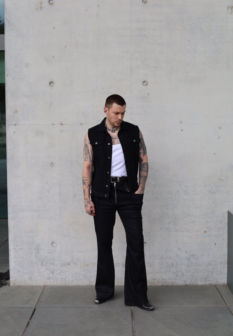Μαύρο τζάκετ από τζιν πάνω από λευκό tank top, συνδυασμένο με μαύρα φαρδιά παντελόνια και ασημένια αξεσουάρ. Ορατά τα τατουάζ στους βραχίονες. Απλή σκηνή με φόντο από σκυρόδεμα.